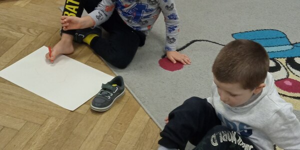 chłopcy siedząc na dywanie rysują stopami po kartce.jpg