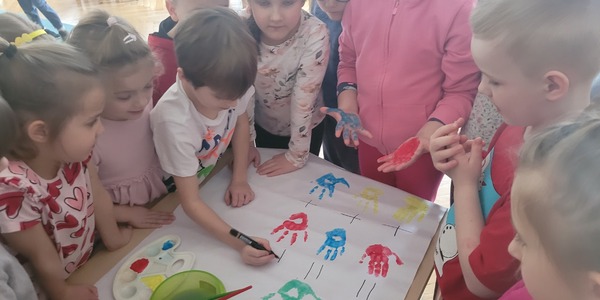 grupa dzieci stoi wokół arkusza papieru z odbitymi na nim dłońmi w różnych kolorach.jpg